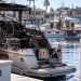 Close-up of a charred boat in a Huntington Harbour residential slip, with firefighters and harbor vessels blurred in the background.