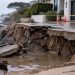 Close-up of a collapsed coastal driveway with exposed earth and standing water near a residential property, illustrating damage caused by coastal erosion.
