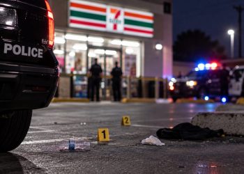 Close-up of a convenience store parking lot with scattered debris, blurred emergency vehicles and police activity in the background, illustrating a shooting in San Bernardino.