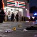 Close-up of a convenience store parking lot with scattered debris, blurred emergency vehicles and police activity in the background, illustrating a shooting in San Bernardino.