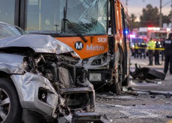 Close-up of a damaged SUV and Metro bus at a Southern California crash scene, with emergency vehicles and investigators blurred in the background.