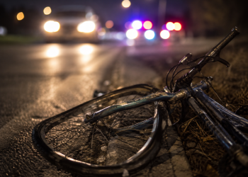 Close-up of a damaged bicycle lying near a dark roadway with blurred headlights and emergency lights in the background, representing a fatal hit-and-run investigation.