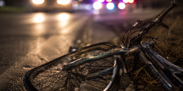 Close-up of a damaged bicycle lying near a dark roadway with blurred headlights and emergency lights in the background, representing a fatal hit-and-run investigation.