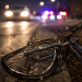 Close-up of a damaged bicycle lying near a dark roadway with blurred headlights and emergency lights in the background, representing a fatal hit-and-run investigation.