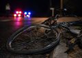 Close-up of a damaged bicycle wheel near the edge of a dark roadway, with a blurred person standing beside distant emergency lights in the background.