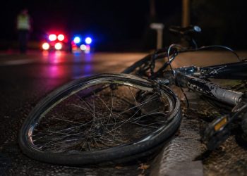 Close-up of a damaged bicycle wheel near the edge of a dark roadway, with a blurred person standing beside distant emergency lights in the background.