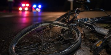 Close-up of a damaged bicycle wheel near the edge of a dark roadway, with a blurred person standing beside distant emergency lights in the background.