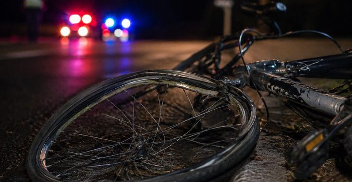 Close-up of a damaged bicycle wheel near the edge of a dark roadway, with a blurred person standing beside distant emergency lights in the background.