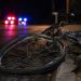 Close-up of a damaged bicycle wheel near the edge of a dark roadway, with a blurred person standing beside distant emergency lights in the background.