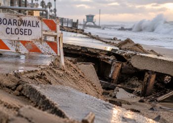 Close-up of a damaged coastal boardwalk and eroded sand berm with large waves in the background, illustrating severe surf damage on the Long Beach Peninsula.