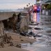 Close-up of a damaged coastal seawall with sand and floodwater along a residential walkway, with blurred emergency lights in the background.