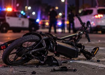 Close-up of a damaged electric bicycle on a Los Angeles roadway at night, with blurred emergency lights and police activity in the background, illustrating a fatal e-bike crash in Arleta.