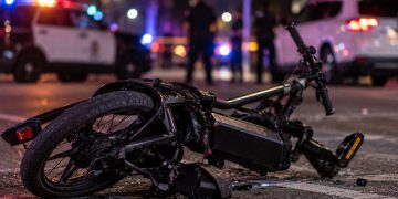 Close-up of a damaged electric bicycle on a Los Angeles roadway at night, with blurred emergency lights and police activity in the background, illustrating a fatal e-bike crash in Arleta.