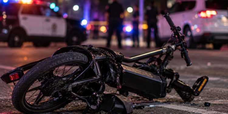 Close-up of a damaged electric bicycle on a Los Angeles roadway at night, with blurred emergency lights and police activity in the background, illustrating a fatal e-bike crash in Arleta.