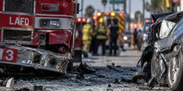 Close-up of a damaged fire apparatus and shattered vehicle glass on a city street, with emergency responders blurred in the background after a multi-vehicle crash in South Los Angeles.
