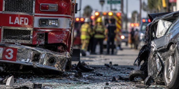 Close-up of a damaged fire apparatus and shattered vehicle glass on a city street, with emergency responders blurred in the background after a multi-vehicle crash in South Los Angeles.