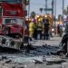 Close-up of a damaged fire apparatus and shattered vehicle glass on a city street, with emergency responders blurred in the background after a multi-vehicle crash in South Los Angeles.