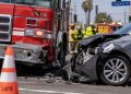 Close-up of a damaged firetruck and civilian vehicle at a South Los Angeles intersection, with traffic cones and blurred emergency responders in the background.