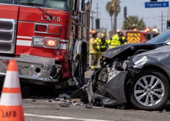 Close-up of a damaged firetruck and civilian vehicle at a South Los Angeles intersection, with traffic cones and blurred emergency responders in the background.