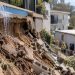 Close-up of a damaged hillside and retaining wall beneath a residential pool, with blurred emergency crews and apartment buildings in the background.
