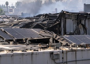 Close-up of a damaged industrial warehouse roof with solar panels and smoke haze in the background, illustrating litigation over the Boyle Heights warehouse fire.