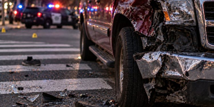 Close-up of a damaged pickup truck and roadway debris near a pedestrian crossing, with blurred emergency lights in the background, illustrating a hit-and-run investigation in Riverside.