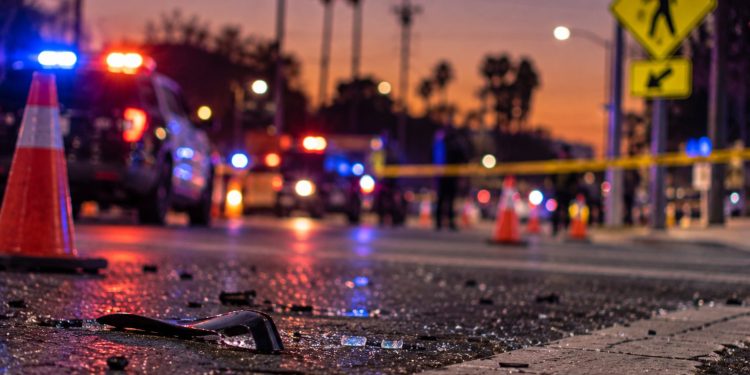 Close-up of a damaged roadway at dawn with blurred emergency lights, traffic cones and a pedestrian crossing sign in the background, illustrating a fatal hit-and-run in the San Fernando Valley.