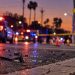 Close-up of a damaged roadway at dawn with blurred emergency lights, traffic cones and a pedestrian crossing sign in the background, illustrating a fatal hit-and-run in the San Fernando Valley.