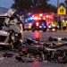 Close-up of a damaged silver Lexus and scattered electric scooter debris at a Riverside intersection, with emergency crews and a school-area street blurred in the background.