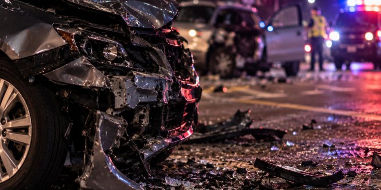 Close-up of a damaged vehicle and roadway debris at night, with blurred emergency lights in the background, illustrating a fatal two-vehicle crash investigation in Encino.