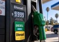 Close-up of a diesel fuel pump displaying $9.999 per gallon, with a green diesel nozzle in sharp focus and a blurred gas station in the background.