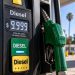 Close-up of a diesel fuel pump displaying $9.999 per gallon, with a green diesel nozzle in sharp focus and a blurred gas station in the background.