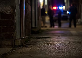 Close-up of a dimly lit alley entrance and pavement at night, with blurred investigators and emergency lights visible farther in the background.