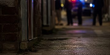 Close-up of a dimly lit alley entrance and pavement at night, with blurred investigators and emergency lights visible farther in the background.