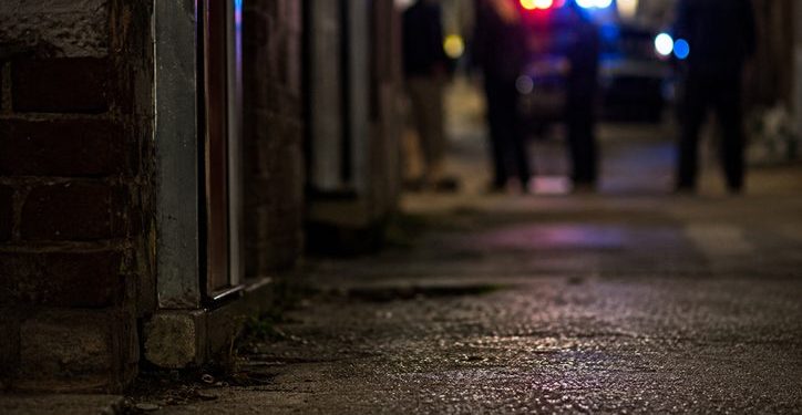 Close-up of a dimly lit alley entrance and pavement at night, with blurred investigators and emergency lights visible farther in the background.