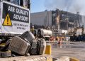 Close-up of a dusty respirator beside an air-quality warning sign near a damaged warehouse, illustrating lingering health concerns after the Lineage fire.