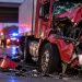 Close-up of a heavily damaged red semi-truck cab with shattered glass and roadway debris, with blurred emergency lights in the background, illustrating a big rig crash investigation.