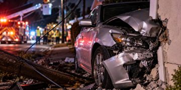 Close-up of a heavily damaged silver sedan against a residential property, with utility wires, emergency crews and a blocked Mission Hills street blurred in the background.