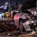 Close-up of a heavily damaged silver sedan against a residential property, with utility wires, emergency crews and a blocked Mission Hills street blurred in the background.