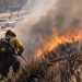 Close-up of a hillside brush fire burning near a freeway, with heavy smoke and firefighters working in the background, illustrating the Getty Fire response in Los Angeles.