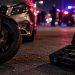 Close-up of a motorcycle beside a damaged vehicle at night, with blurred emergency lights and a firearm case in the background, illustrating a road rage investigation in Camarillo.