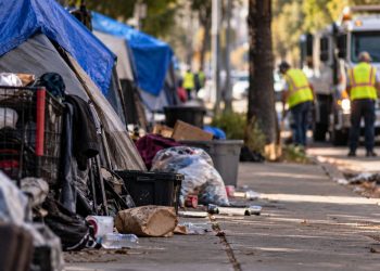 Close-up of a neighborhood sidewalk with tents, discarded belongings and scattered trash, with a residential street and blurred city cleanup activity in the background.