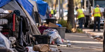 Close-up of a neighborhood sidewalk with tents, discarded belongings and scattered trash, with a residential street and blurred city cleanup activity in the background.