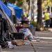 Close-up of a neighborhood sidewalk with tents, discarded belongings and scattered trash, with a residential street and blurred city cleanup activity in the background.