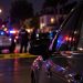 Close-up of a parked vehicle at night with blurred emergency lights, police activity and residential homes in the background, illustrating a fatal shooting in Granada Hills.