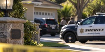 Close-up of a quiet Newbury Park residential street with a home entrance and blurred sheriff's vehicles in the background, illustrating a death investigation.