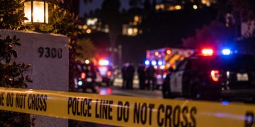 Close-up of a residential street at night with blurred police lights, crime-scene tape and emergency responders in the background, illustrating an officer-involved shooting in the Hollywood Hills.