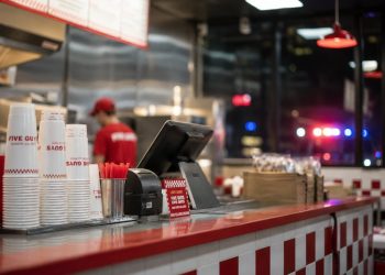 Close-up of a restaurant counter and food-service area, with a blurred employee in the background and distant emergency lights visible outside.
