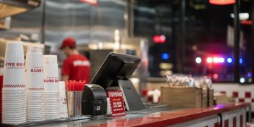 Close-up of a restaurant counter and food-service area, with a blurred employee in the background and distant emergency lights visible outside.