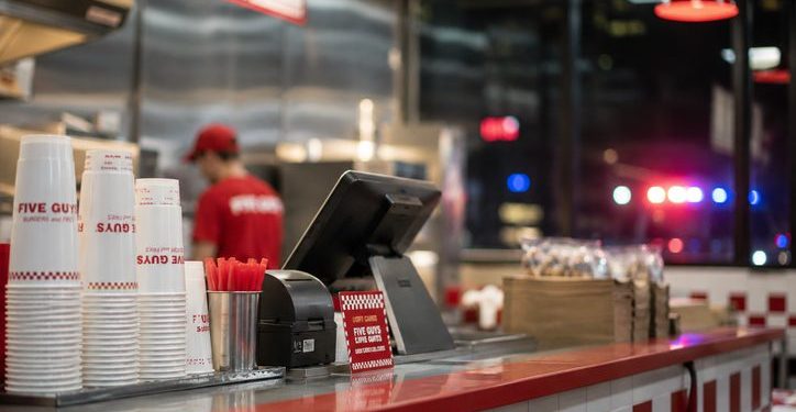 Close-up of a restaurant counter and food-service area, with a blurred employee in the background and distant emergency lights visible outside.